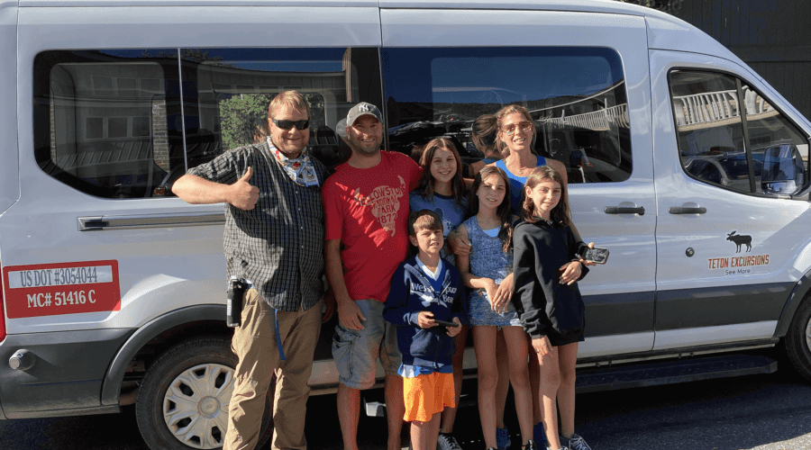 A guide poses in front of a Teton Excursions van with a young family. A Teton Excursions raised roof van in front of the Teton Mountains. Waiting for the guests at Teton Village. Taken on a Teton Excursions summer tour of Grand Teton National Park.