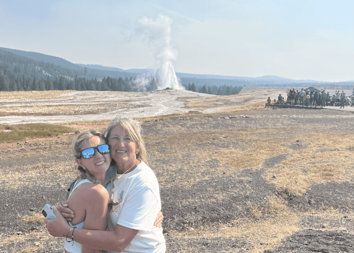 A mother and daughter pose together for a photo as Old Faithful geyser erupts in the background. Taken on a Teton Excursions tour of Yellowstone National Park.